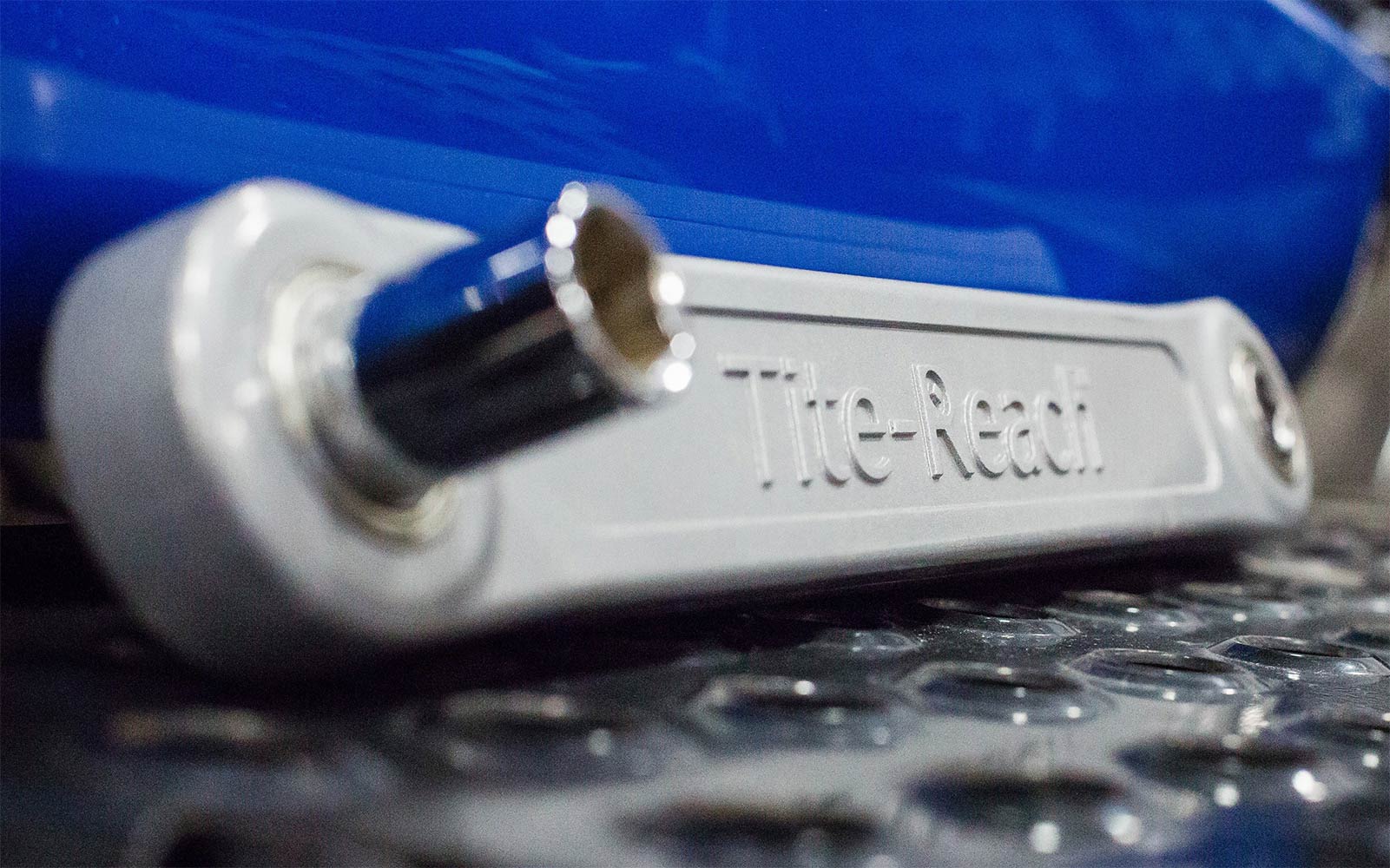 A Tite-Reach extension wrench, in a light gray color, lies on a metallic, textured surface with a blue background.