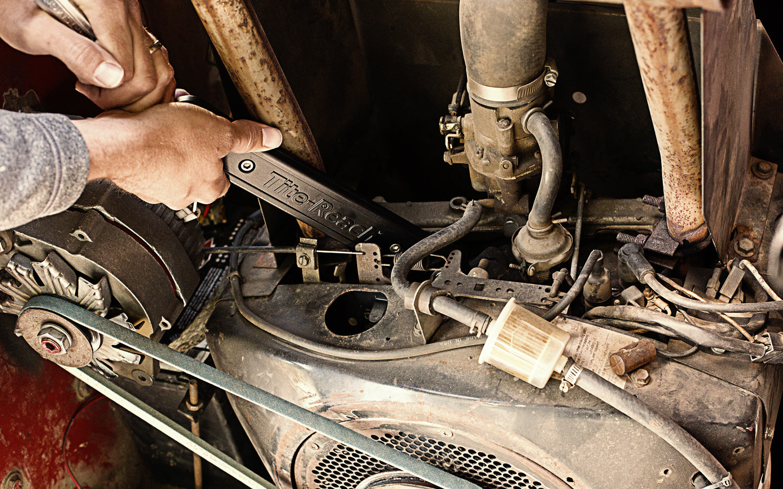 A pair of hands uses a black Tite-Reach wrench on an engine surrounded by belts, hoses, and mechanical components.